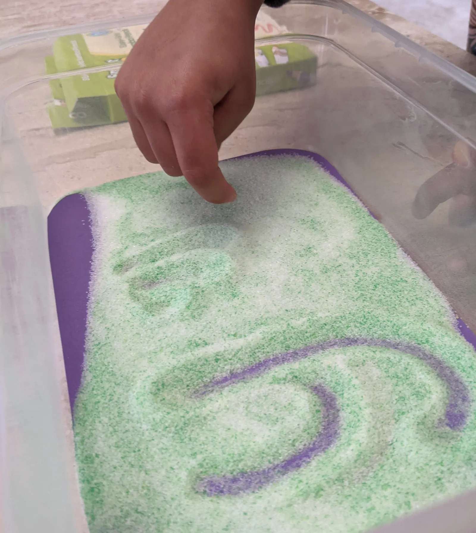 A child's finger drawing a letter in a tray of coloured salt